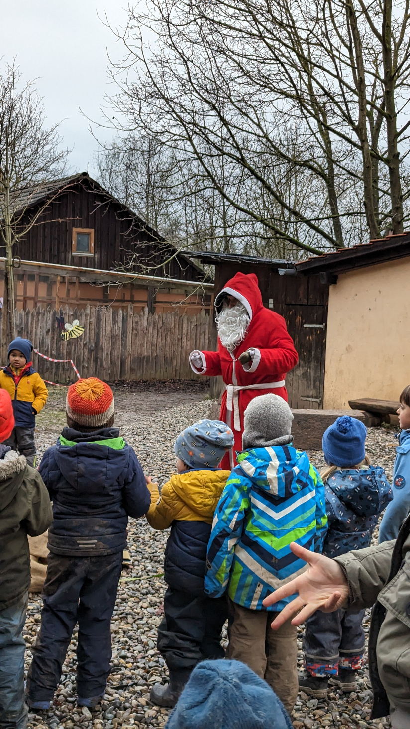 Kinder in Winterkleidung schauen zu einem als Nikolaus verkleideten Erwachsenen auf, der in einem offenen Hof vor einem Holzhaus steht.
