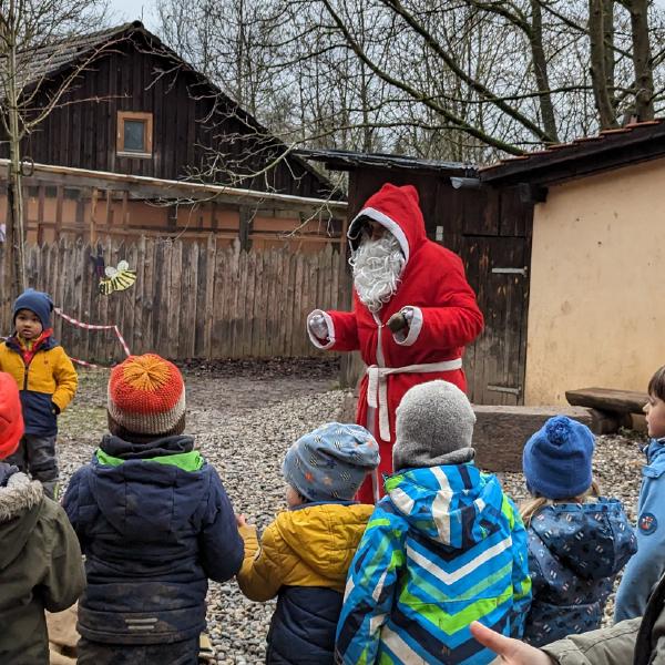 Kinder in Winterkleidung schauen zu einem als Nikolaus verkleideten Erwachsenen auf, der in einem offenen Hof vor einem Holzhaus steht.